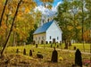 A white church with a steeple stands in a small graveyard surrounded by trees with autumn foliage under a partly cloudy sky.