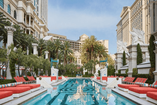 A luxurious pool surrounded by red lounge chairs, palm trees, and ornate classical buildings under a clear sky.