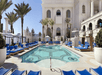 Luxurious pool area with blue lounge chairs and cabanas, surrounded by palm trees and a grand building in the background.