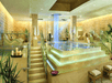 Luxurious spa interior featuring a central multi-tiered pool with cascading waterfall, surrounded by lounge chairs, plants, and soft lighting.