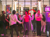 A group of people dance and socialize indoors at night, illuminated by colorful lights, with large windows and art displays in the background.