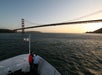 Two people stand at the bow of a boat looking out at the Golden Gate Bridge over the water at sunset.