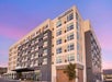 A modern multi-story hotel building with "CAMBRiA" signage, featuring large windows and a mix of beige and dark panels, set against a twilight sky.