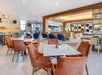 Modern lounge with marble tables, orange and blue chairs, wooden accents, bookshelves, and decorative wall art. Bright lighting and spacious layout.