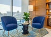 Two blue chairs face each other with a round table and potted plant between them, set on a patterned rug. A tall lamp stands nearby, with sunlight streaming through a large window.
