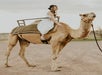 A woman in a white dress sits on a camel in a desert setting under a cloudy sky.