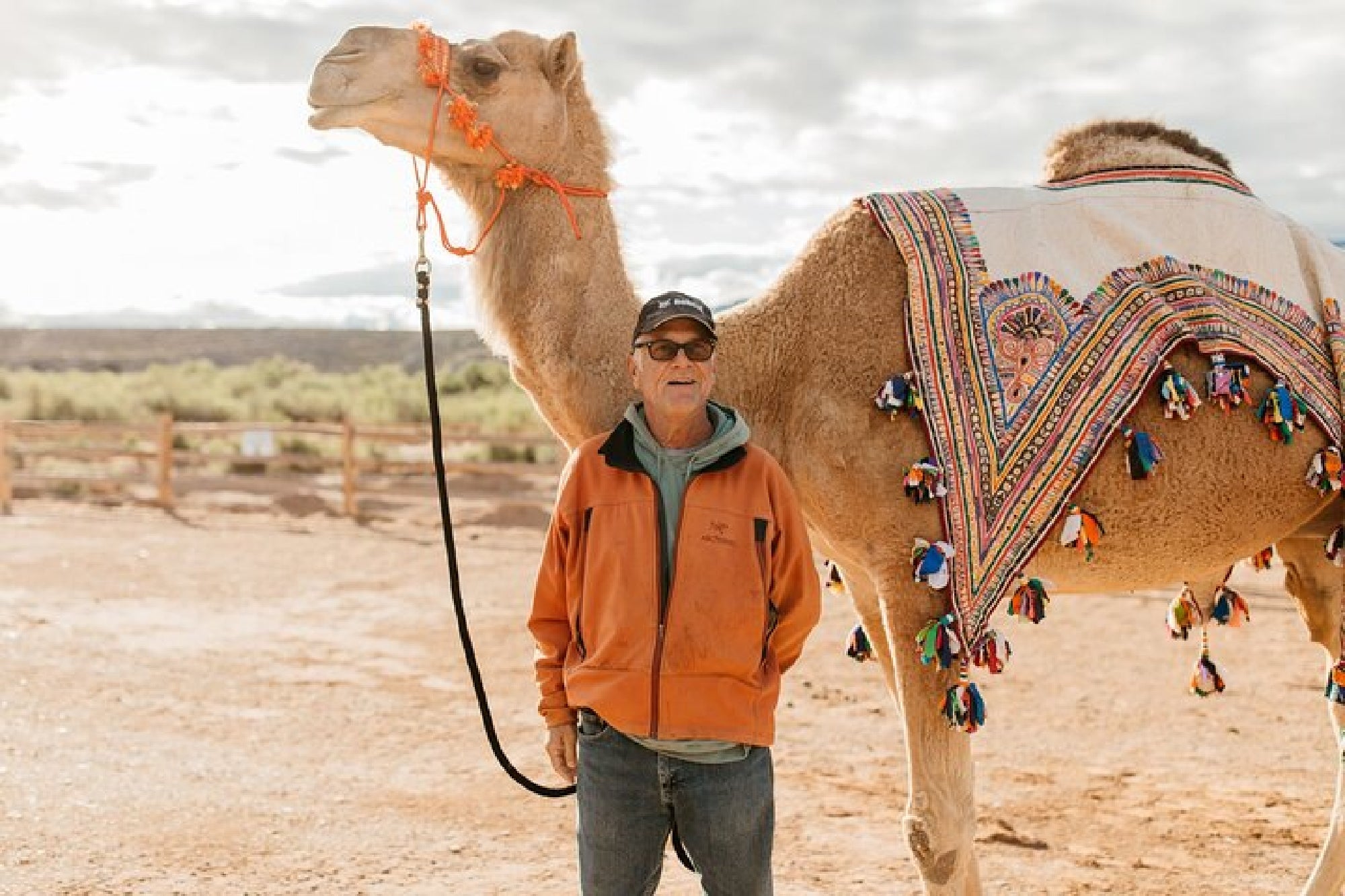 Man standing next to a camel wearing a decorated blanket in a desert setting, under a cloudy sky.