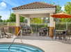 Outdoor pool area with a covered patio, tables, chairs, two barbecue grills, and an orange umbrella, surrounded by greenery and a white fence.