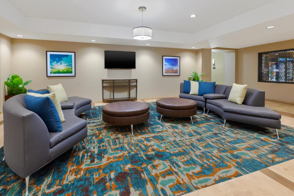 Modern lounge area with gray curved sofas, colorful cushions, round leather ottomans, and an abstract-patterned rug. Walls feature framed art; a TV is mounted above a console table.
