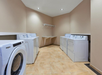 Laundry room with tiled floor, featuring four white machines: two washers and two dryers. A wall-mounted shelf and fluorescent lighting are present. Walls are painted beige.