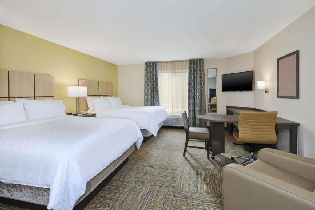 Hotel room with two large beds, a desk with chair, a wall-mounted TV, and a window with curtains. Floor is carpeted, walls are light-colored, and room is well-lit.