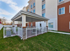 Outdoor patio area with a covered gazebo, surrounded by a white fence. The area has a barbecue grill, seating, and is adjacent to a multi-story building with several windows.