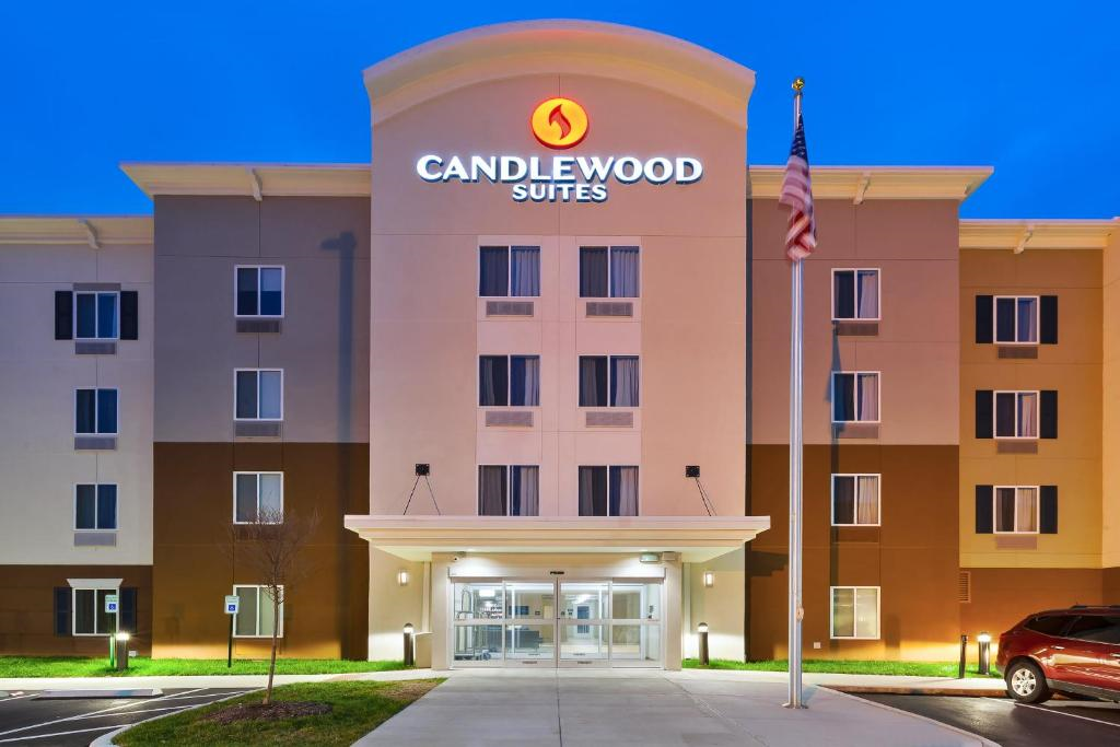Exterior of Candlewood Suites hotel at dusk with illuminated sign. The building is four stories tall with a central entrance and an American flag on a pole in front.