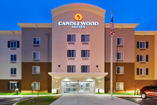 Exterior of Candlewood Suites hotel at dusk with illuminated sign. The building is four stories tall with a central entrance and an American flag on a pole in front.