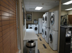 A laundry room with multiple washing machines and dryers, a trash can, and a shelf with towels.