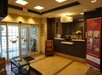 Hotel lobby with a reception desk, a leather chair, and a promotional banner. Glass doors lead outside, and a ceiling light fixture illuminates the space.