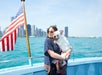 A woman wearing sunglasses holds a French bulldog on a boat, with an American flag and city skyline visible in the background.