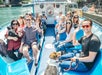 A group of people and a dog sit on a blue and white boat, smiling and waving at the camera as the boat travels along a city waterway on a sunny day.