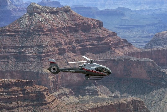 A Maverick Airbus helicopter flying by Point Imperial and Marble Canyon with the rocky formations in the background on a sunny day.