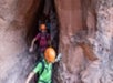 Three people wearing helmets navigate a narrow rocky canyon, each using their arms for support against the walls.