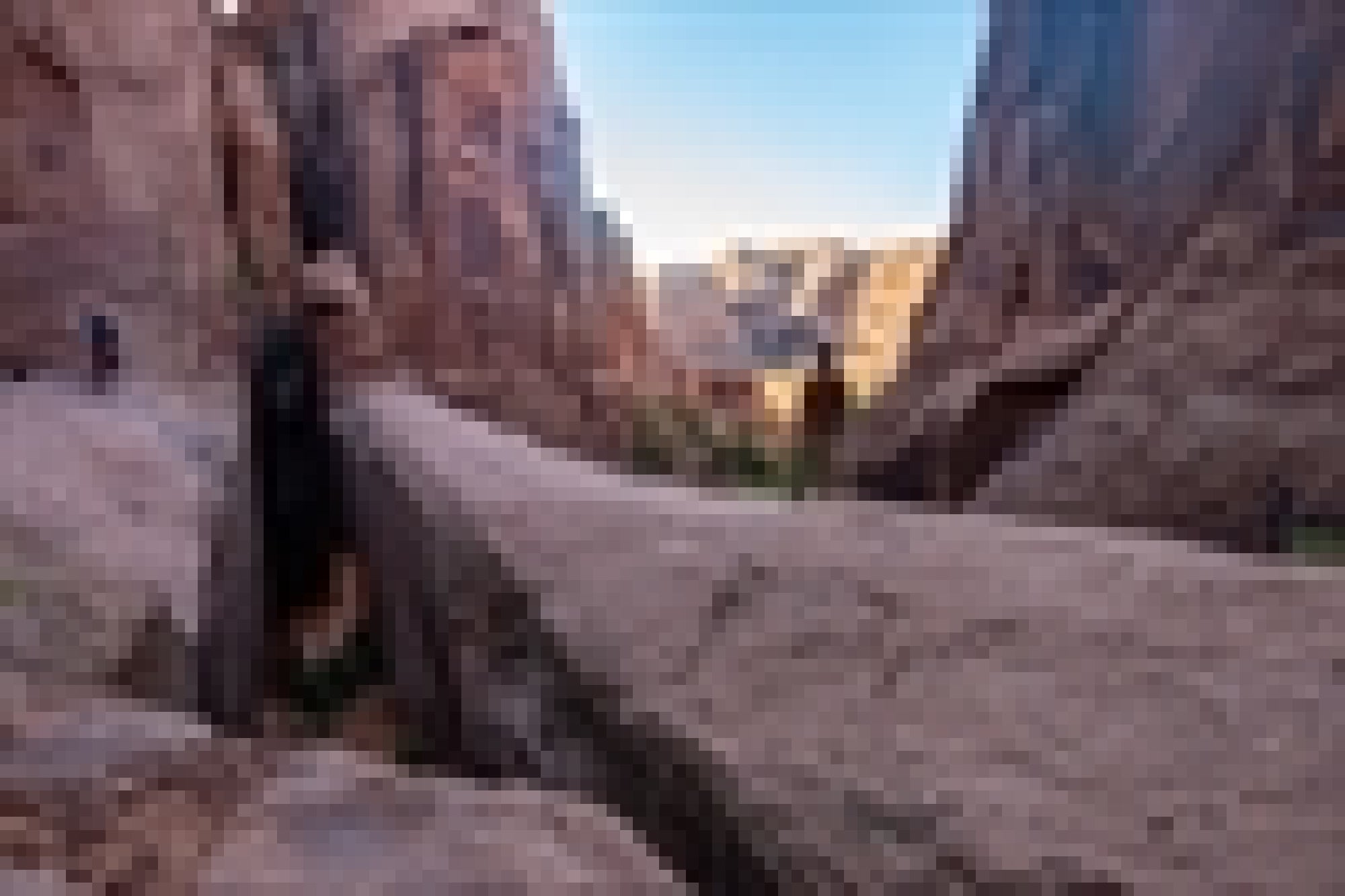 People walk along a narrow rock ledge between tall, rugged cliffs under a clear blue sky.