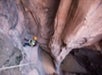 A person in climbing gear ascends a steep rock face in a narrow canyon, wearing a helmet and harness. The surrounding rock is brown and textured.