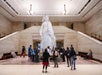 A group of people stand around a large white statue in the center of a spacious, well-lit lobby with beige stone walls and staircases on both sides.