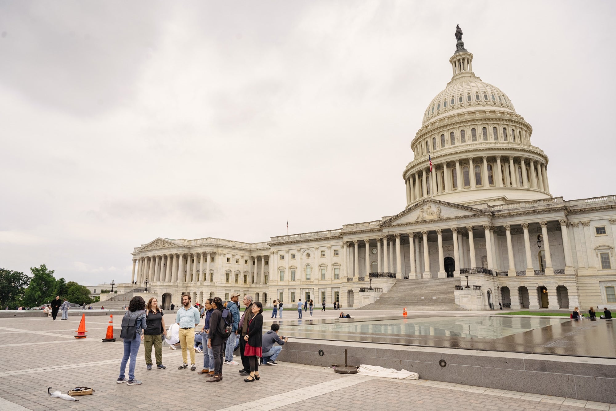 A small group of people stands near the reflecting pool outside the United States Capitol building on a cloudy day.