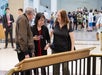 Three adults stand and converse near a wooden railing inside a busy building with people in the background.