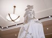 White marble statue wearing a headdress, viewed from below indoors with ceiling lights and a round modern chandelier visible above.