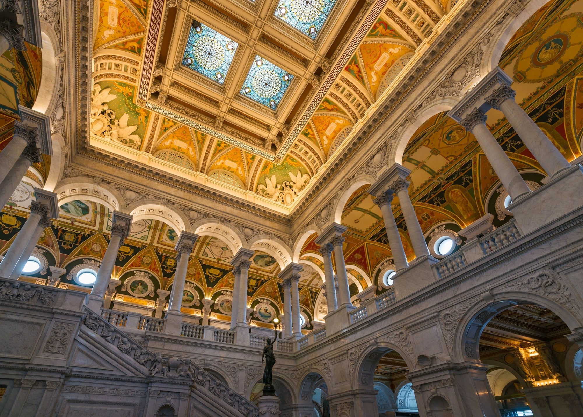 Interior of a grand hall with ornate columns, marble staircases, arched windows, and a decorated ceiling with stained glass and murals.