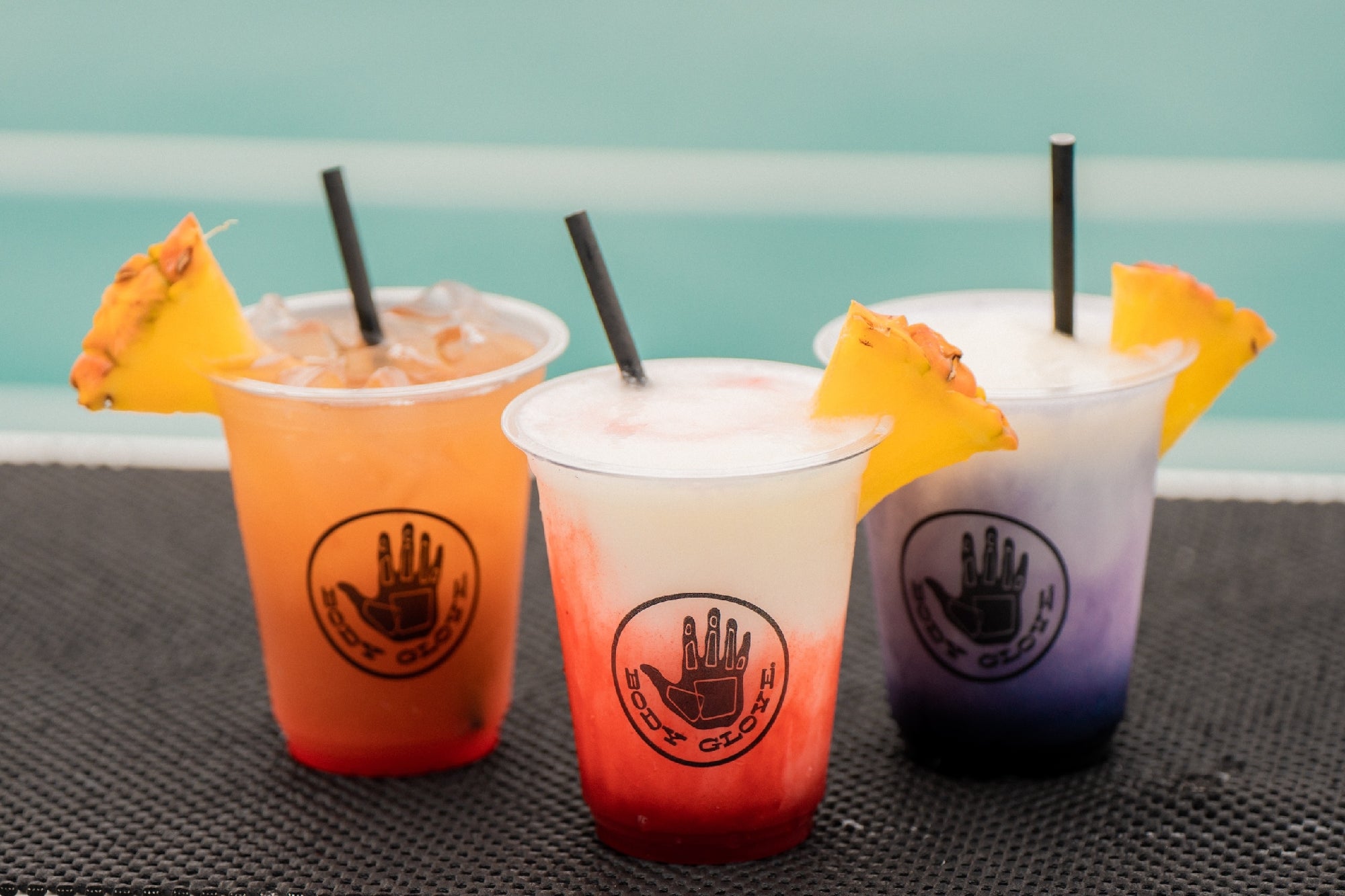 Three colorful tropical drinks in orange, red and blue colors, served in clear cups, each garnished with a slice of pineapple and featuring distinct layered colors, set against a backdrop of light blue ocean water.