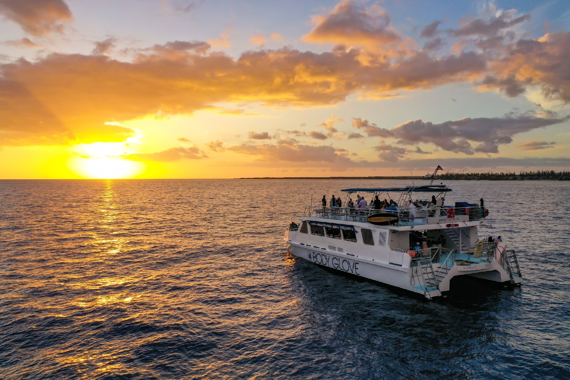 A catamaran named "Body Glove" cruising on the ocean during a vibrant sunset, with people on board enjoying the view.