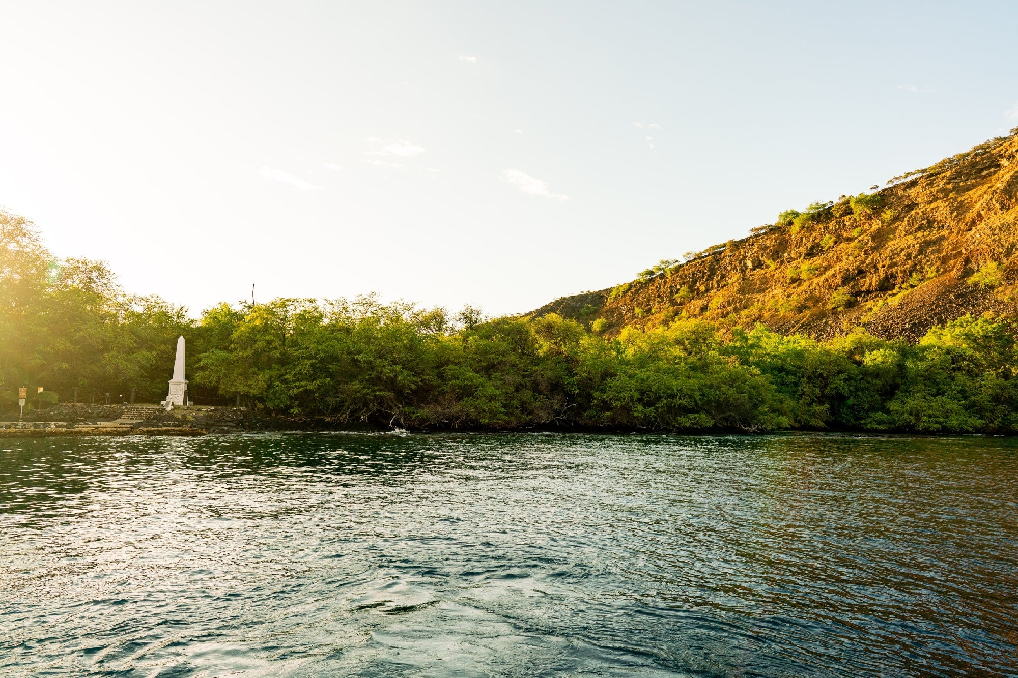 A serene landscape with a body of water in the foreground, framed by lush greenery and a hillside, featuring a prominent white monument standing peacefully along the shore.