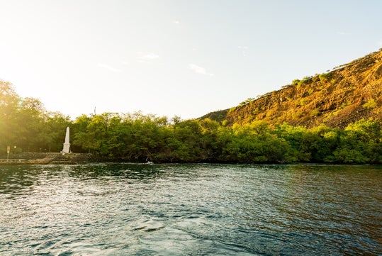 A serene landscape with a body of water in the foreground, framed by lush greenery and a hillside, featuring a prominent white monument standing peacefully along the shore.