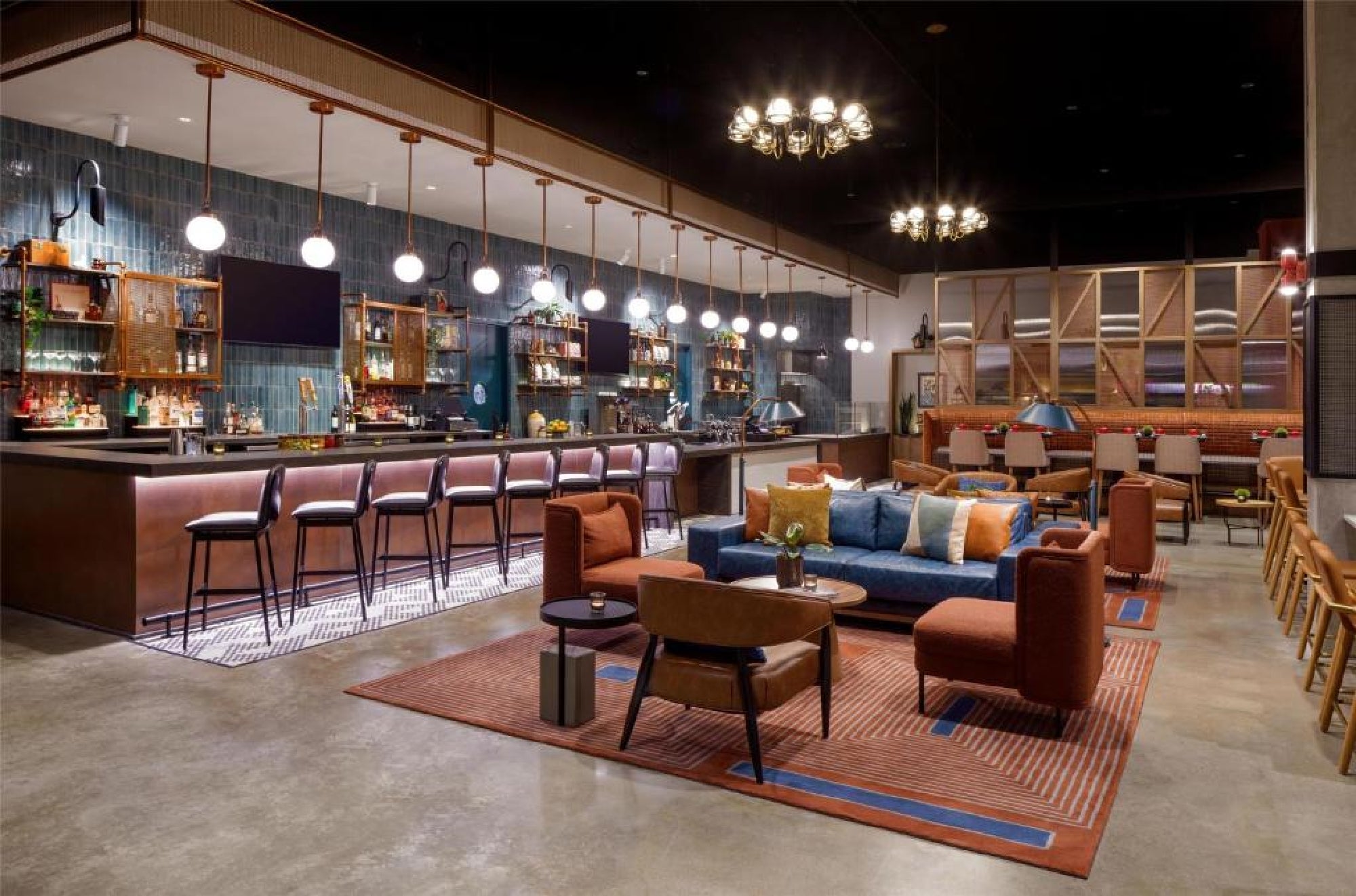 Modern lounge with a bar, upholstered sofas and chairs, a striped rug, pendant lighting, and shelves with bottles behind the bar.