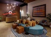Modern hotel lobby with a curved blue sofa, brown ottomans, striped rug, plants, and a reception desk. Geometric lighting and contemporary wall art decorate the space.