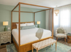 A bedroom with a wooden canopy bed, white bedding, green accent pillows, two green lamps on nightstands, a woven bench, and a patterned chair on a blue rug.
