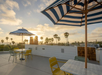 Rooftop patio with striped umbrellas, colorful chairs, and tables overlooking palm trees at sunset.