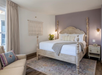 A bedroom with a four-poster bed, purple accent wall, gray throw, pillows, a nightstand, sofa, and wall hanging. There's a large window with curtains and a potted plant in the corner.