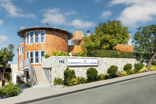A round wooden building with large windows and stone walls, labeled "Cardiff by the Sea Lodge." Surrounded by trees and plants on a sloped street.