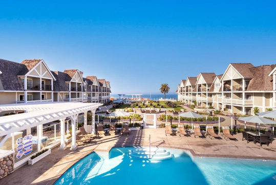 A resort with multiple two-story buildings, a swimming pool in the foreground, palm trees, and a view of the ocean in the background under a clear blue sky.