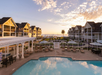 A resort with a central swimming pool, surrounded by buildings with gabled roofs. Outdoor seating and gardens lead to a view of the ocean and a sunset sky.