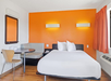Hotel room with an orange accent wall, white bed, wall-mounted lights, a small round table with chairs, and a window with orange curtains.