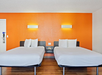 Two double beds with white linens in a hotel room with an orange accent wall, wooden floor, and minimalistic decor.