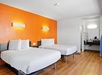 Hotel room with two double beds, orange accent wall, wooden flooring, wall-mounted lights, and an open vanity area with a sink and mirror.