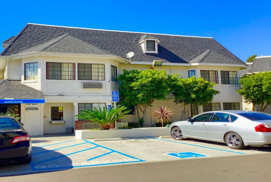 A two-story building with a dark roof and white walls, featuring large windows. Two cars are parked in spaces designated for disabled parking.