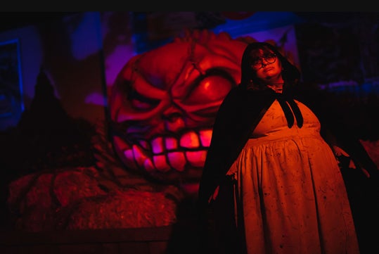 A person in a cape and dress stands in front of a large, menacing pumpkin face decoration, lit by red and orange light.