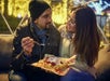 A man and woman sit together on a couch outdoors at night, sharing a plate of food and smiling at each other, with lights in the background.