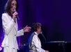 A woman in a white suit sings into a microphone while a man in a white shirt plays the piano and sings on a stage with purple lighting.